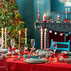 A festive Christmas dinner table decorated with red tablecloths, striped taper candles, crystal wine glasses, Santa-themed napkins, and holiday ornaments, set in a decorated room with a Christmas tree and fireplace.