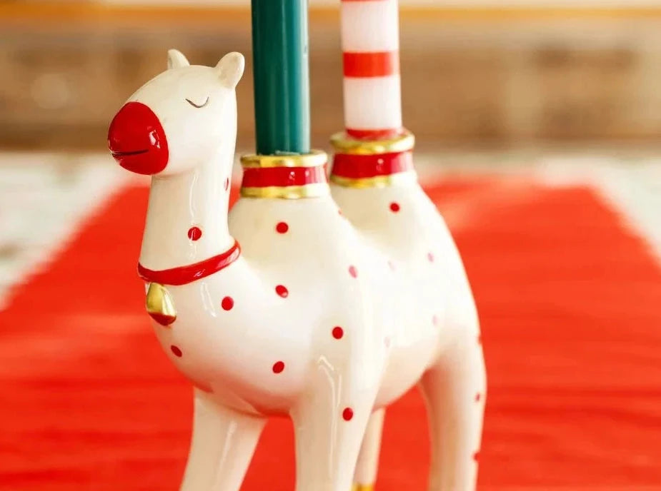 A white ceramic llama-shaped candle holder with red polka dots, gold trim, and gold hooves, holding two candles on a red runner.