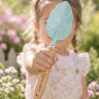 A young girl in a floral dress holds a small leaf-shaped hand trowel with a light blue metal head and a wooden handle engraved with 'Lola's Pretty T.