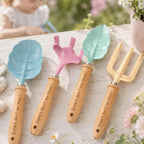 Four engraved wooden-handled garden tools with leaf-shaped heads are arranged on a white wooden surface outdoors. The set includes a trowel in light blue, a small cultivator in pink, a seed scatterer in mint green, and a three-prong garden fork in pale yellow.