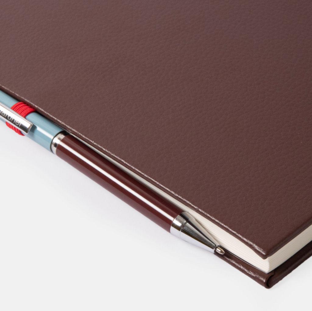 A close-up of a dark brown textured notebook or journal with a matching pen tucked along the edge, photographed on a clean white background.