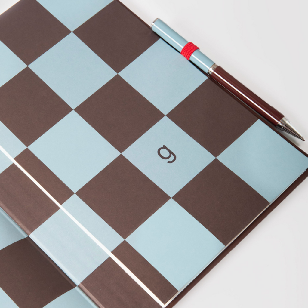 A notebook with a brown and light blue checkerboard cover, featuring a small stylized logo in the center of a light blue square. A silver and dark brown pen with a red band rests diagonally across the top right corner of the notebook on a white background.