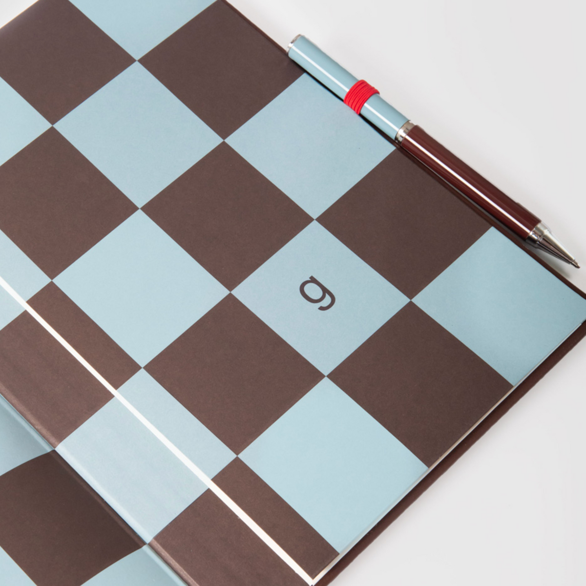 A notebook with a brown and light blue checkerboard cover, featuring a small stylized logo in the center of a light blue square. A silver and dark brown pen with a red band rests diagonally across the top right corner of the notebook on a white background.