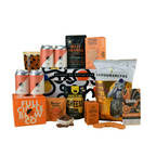 A curated gift basket of snacks, cheese, beer, and coffee arranged on a warm yellow background. Visible products include a bag of Savoursmiths Rugged Country Hilli cheese, a bag of Gnaw Chocolate, a box of Butter Cakes, a can of Billy Franks, a can of Full Circle Brew Co, and a can of Suckler.