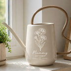 A ceramic watering can with a wavy handle and spout, featuring a white floral illustration and the text 'Mummy September' on its front. It is styled on a windowsill next to a potted green plant and a framed picture.