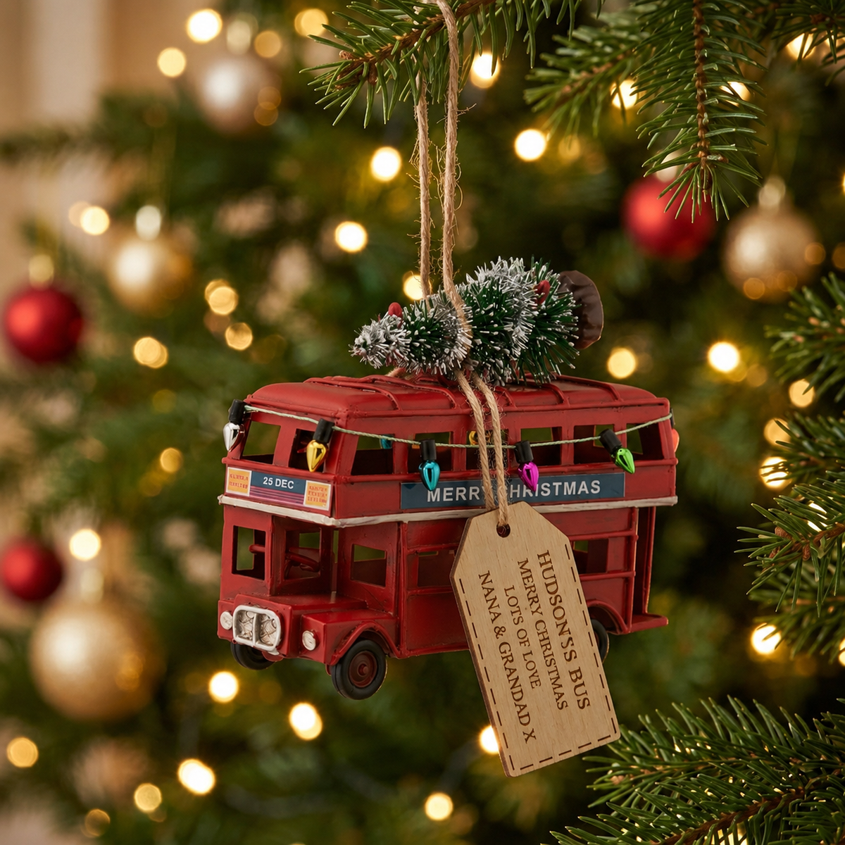 A red double-decker bus Christmas ornament hanging from a tree branch, with a kraft paper tag that reads 'PILGRIM'S RAN' and 'MERRY CHRISTMAS' on the roof, against a blurred Christmas tree background with warm lights.