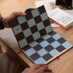 A person holds open a notebook with a checkerboard pattern on the cover, placed on a wooden desk with books and a camera in the background.