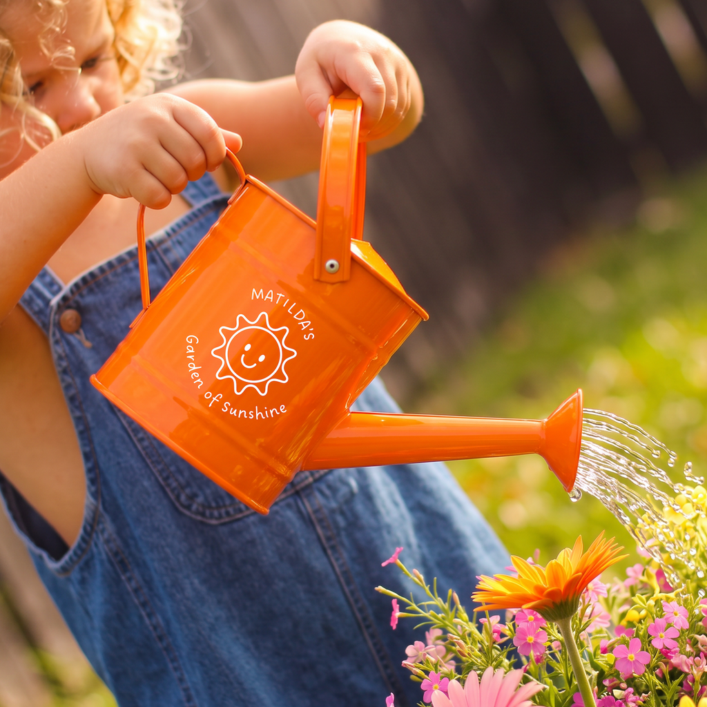 A child in blue denim overalls holds an orange watering can and waters a colorful flower bed outdoors. The can has a white printed logo that reads 'NOT A DAY GARDEN OF SUNSHINE' with a smiling sun graphic.