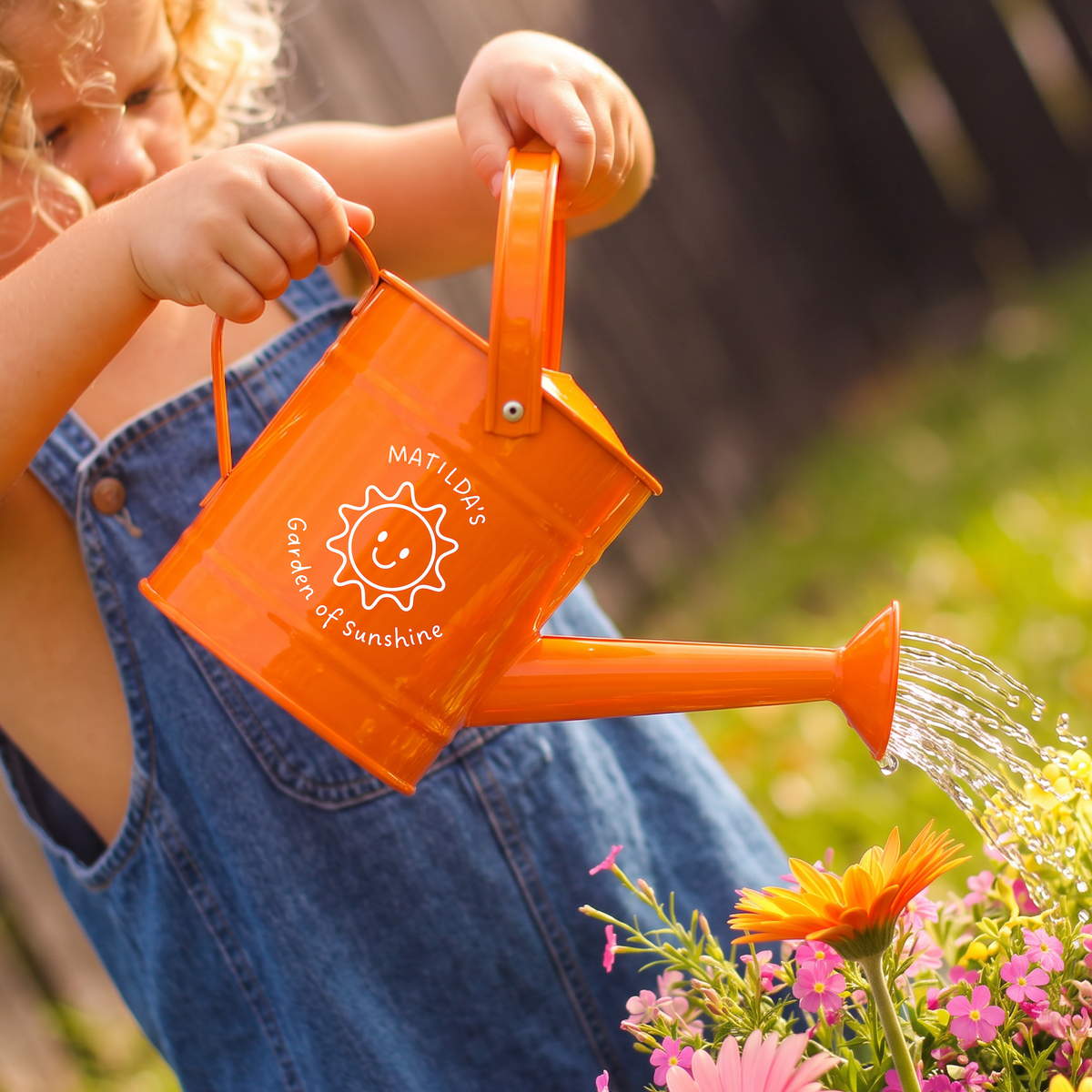 A child in blue denim overalls holds an orange watering can and waters a colorful flower bed outdoors. The can has a white printed logo that reads 'NOT A DAY GARDEN OF SUNSHINE' with a smiling sun graphic.