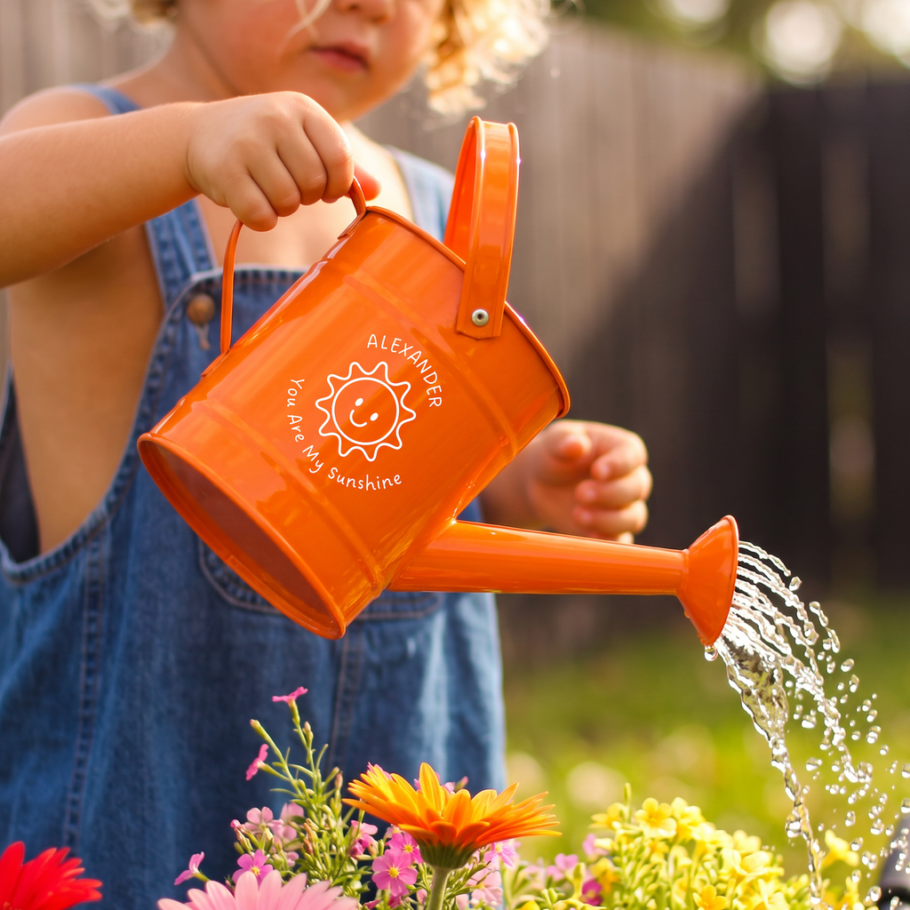 A child in denim overalls holds an orange metal watering can and waters colorful flowers in a garden. The can has a white printed smiley sun design with the text 'ALEXANDER You Are My Sunshine'.