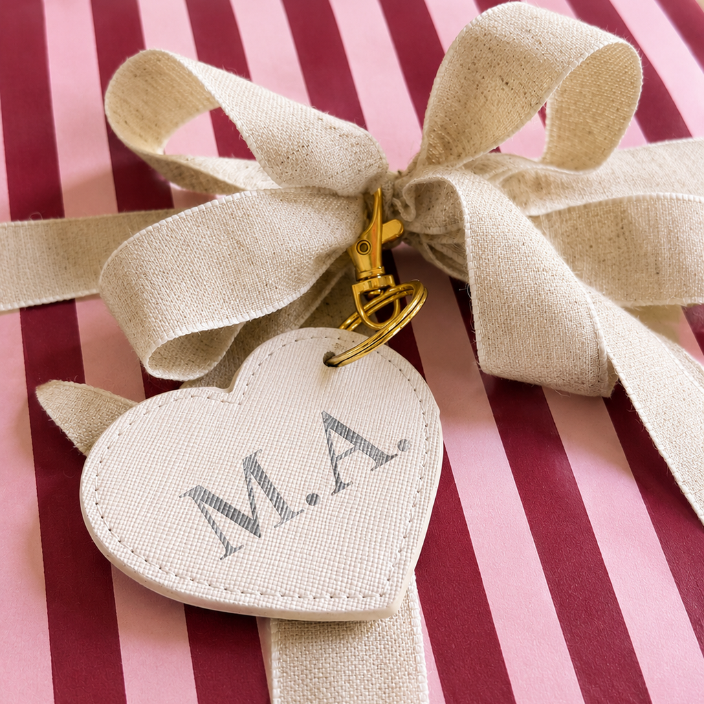 A close-up of a beige ribbon bow with a gold clasp holding a stitched heart-shaped tag printed with the initials 'M.A.