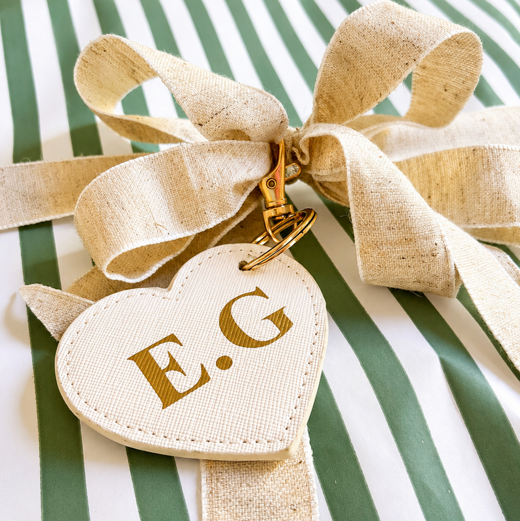 A close-up of a personalized heart-shaped luggage tag made of cream-colored linen fabric with gold embroidered initials 'E.G', tied with a matching cream ribbon on a green and white striped surface.