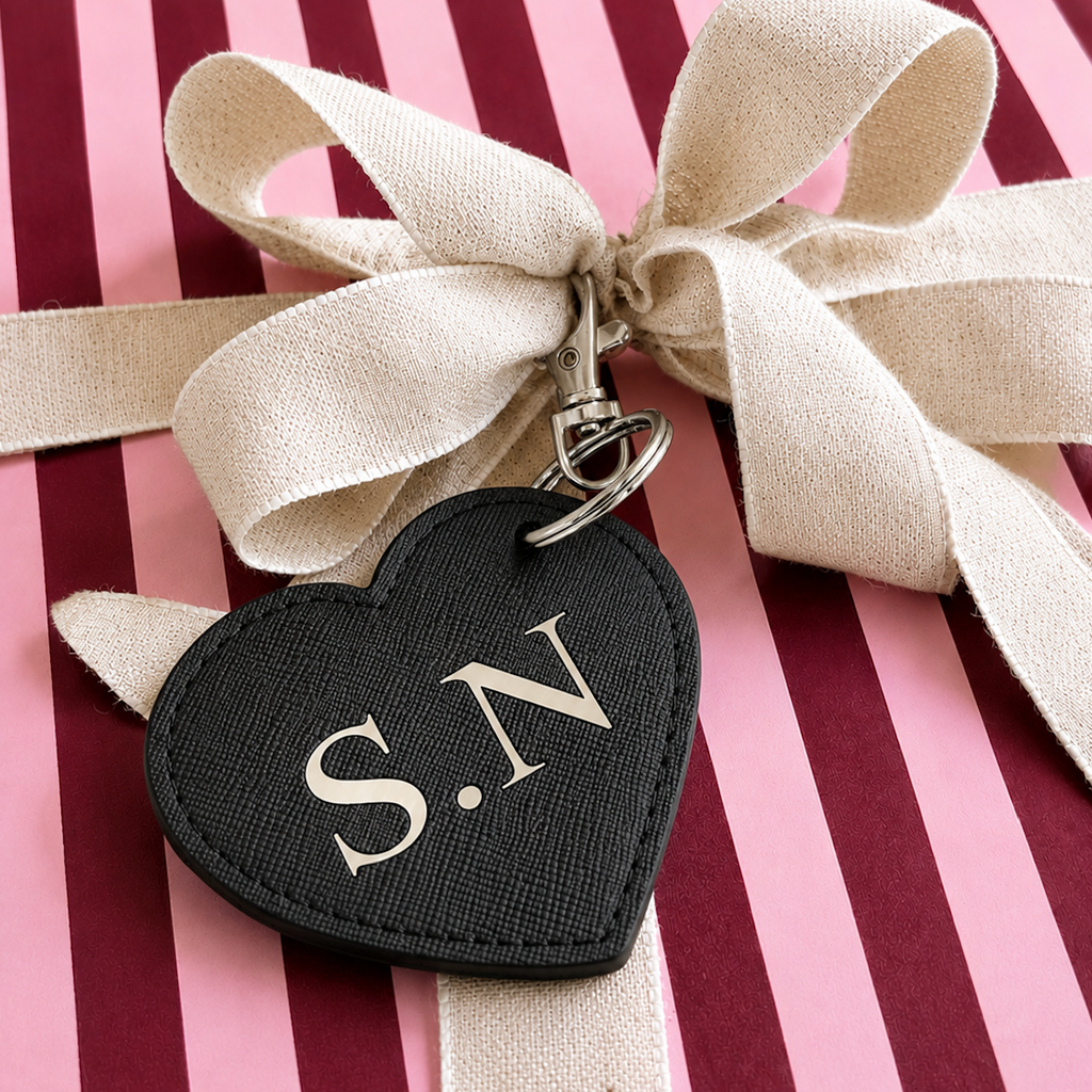 A black heart-shaped keychain or bag charm with silver-tone metal hardware, featuring the initials 'S.N' in a light beige serif font.