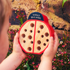 A child holds a ladybug-shaped wooden bug hotel outdoors in front of a stone wall and flowers. The bug hotel has a natural wood body, red shell details, and a dark blue top sign that reads 'SOFA'S BUG HOTEL'.