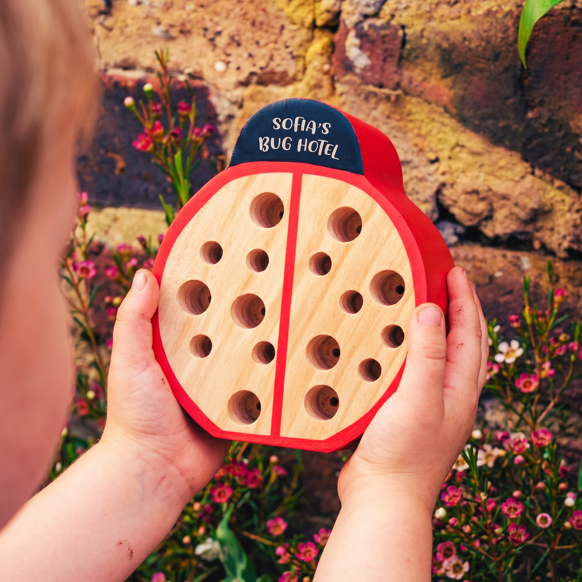 A child holds a ladybug-shaped wooden bug hotel outdoors in front of a stone wall and flowers. The bug hotel has a natural wood body, red shell details, and a dark blue top sign that reads 'SOFA'S BUG HOTEL'.