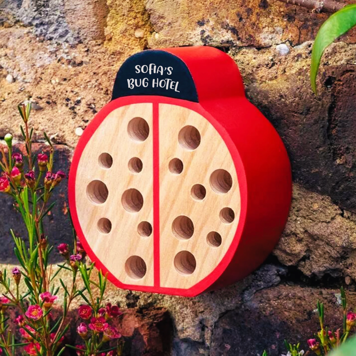 A ladybug-shaped wooden bug hotel with a red shell, light natural wood center, and a small black sign reading 'SOFA'S BUG HOTEL' mounted against a rustic stone wall with flowers nearby.