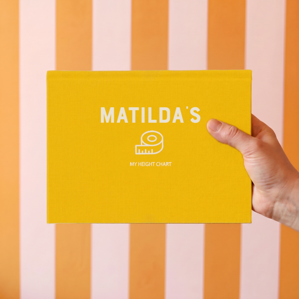 A hand holding a bright yellow height chart book titled 'MATILDA'S MY HEIGHT CHART' against a background of vertical orange and cream stripes.