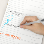 A close-up of a hand holding a clear pen and writing on a lined measurement notebook page with handwritten notes in blue and orange ink.