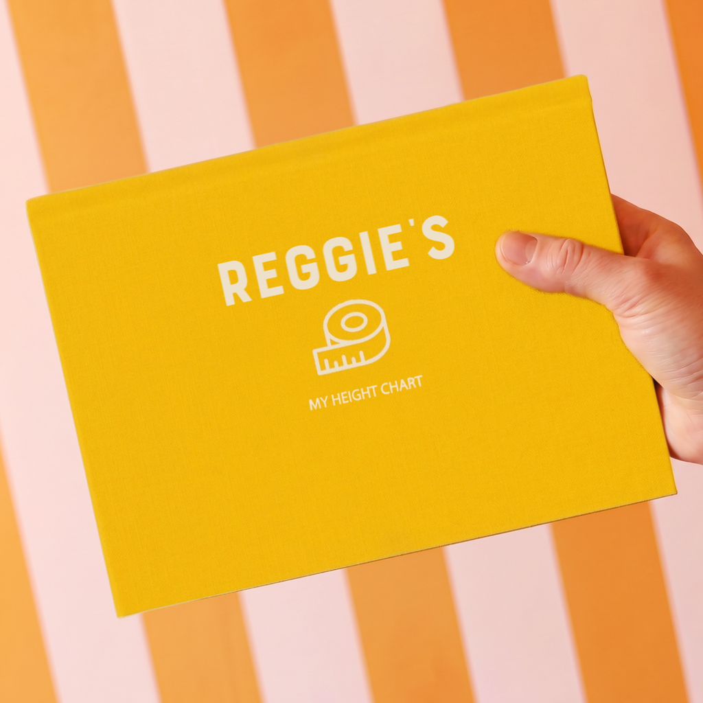 A hand holding a bright yellow personalized height chart book titled 'REGGIE'S MY HEIGHT CHART' against a pink and orange striped background.