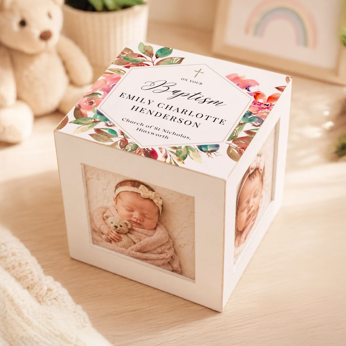 A personalized baby keepsake photo cube box on a light wood surface, with floral artwork around the top lid and two photo windows showing a sleeping newborn baby. The top label reads 'To Our Baptism Emily Charlotte Henderson' with church details below.