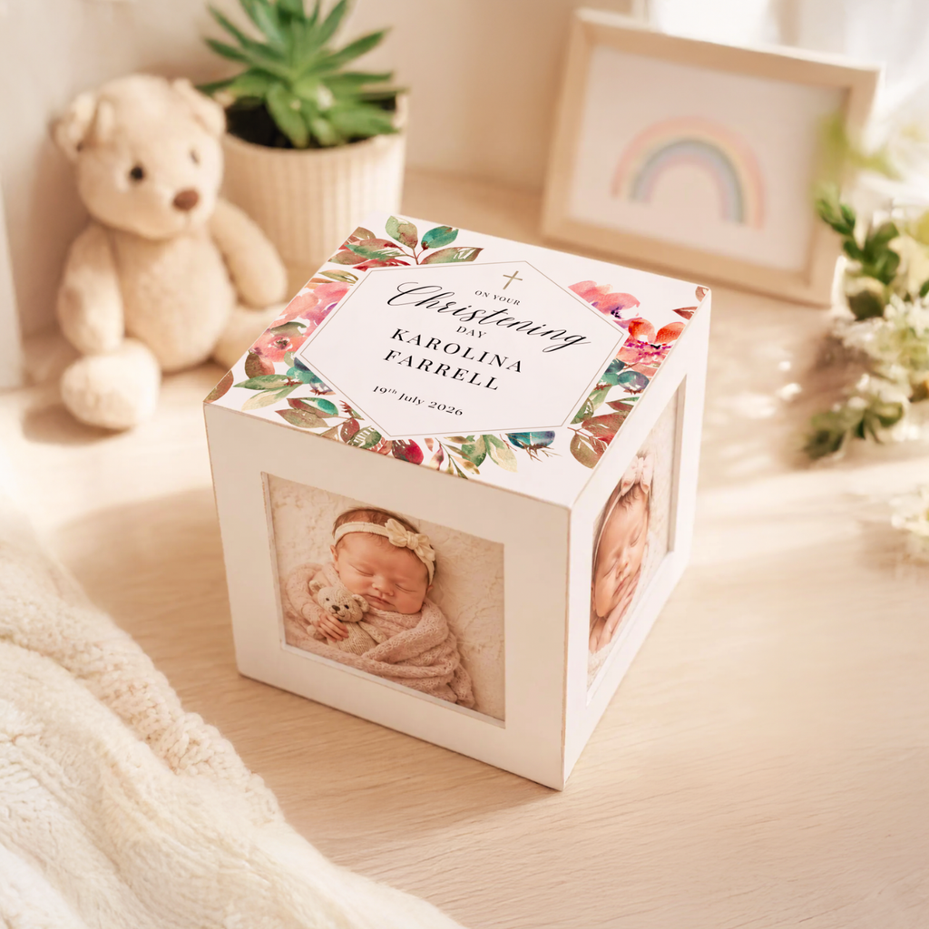 A white wooden keepsake photo cube with floral artwork on the lid and printed baby photos on the sides, styled on a light wood surface with a teddy bear, plant, and framed rainbow in the background.