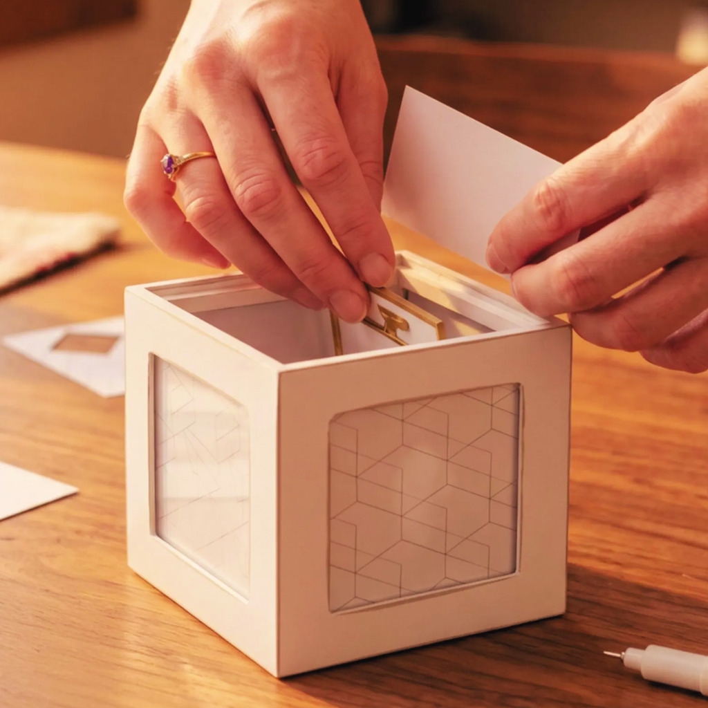 A person's hands are shown inserting a gold card into a white geometric cube box with clear front panels, placed on a wooden table.