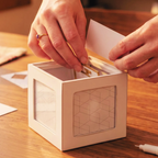 A person's hands are shown inserting a gold card into a white geometric cube box with clear front panels, placed on a wooden table.
