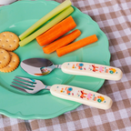 A children's spoon and fork with cream-colored handles decorated with colorful cartoon animals, resting on a mint green plate with carrot sticks, celery, and crackers on a brown-and-white checkered cloth.