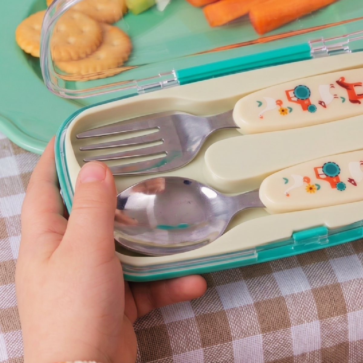 A hand holds an open teal and cream plastic lunchbox utensil case containing a metal fork and spoon with decorative cream handles. A lunchbox with crackers and vegetables is visible in the background on a checkered tablecloth.
