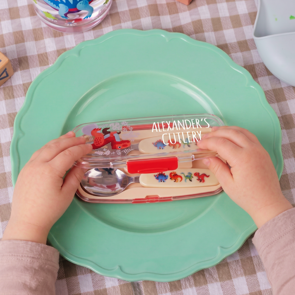 A child's hands holding a clear plastic case of personalized cutlery with dinosaur-themed handles and a spoon, placed on a mint green plate over a checkered tablecloth.