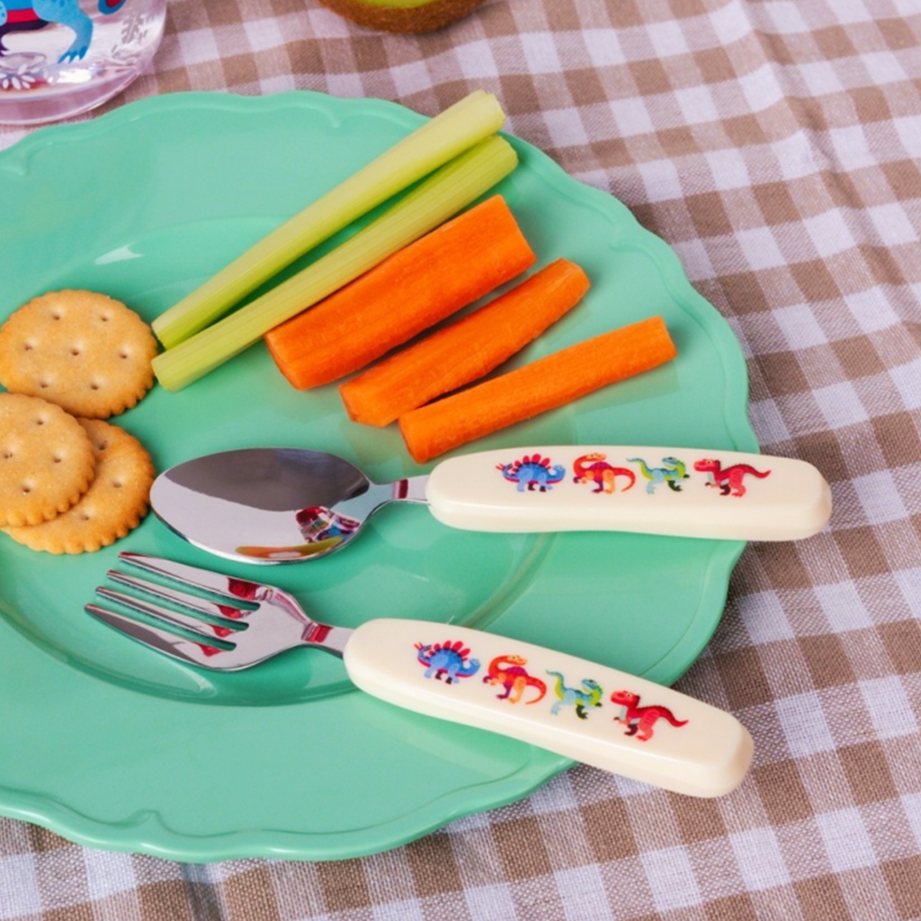 A children's spoon and fork set with cream-colored handles decorated with colorful dinosaur illustrations, shown on a mint green plate with carrots, celery, and crackers on a checkered tablecloth.