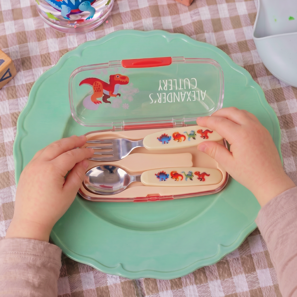 A child's cutlery set in a translucent green case with dinosaur illustrations, held open by two hands over a green plate on a checkered tablecloth.