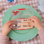A child's cutlery set in a translucent green case with dinosaur illustrations, held open by two hands over a green plate on a checkered tablecloth.