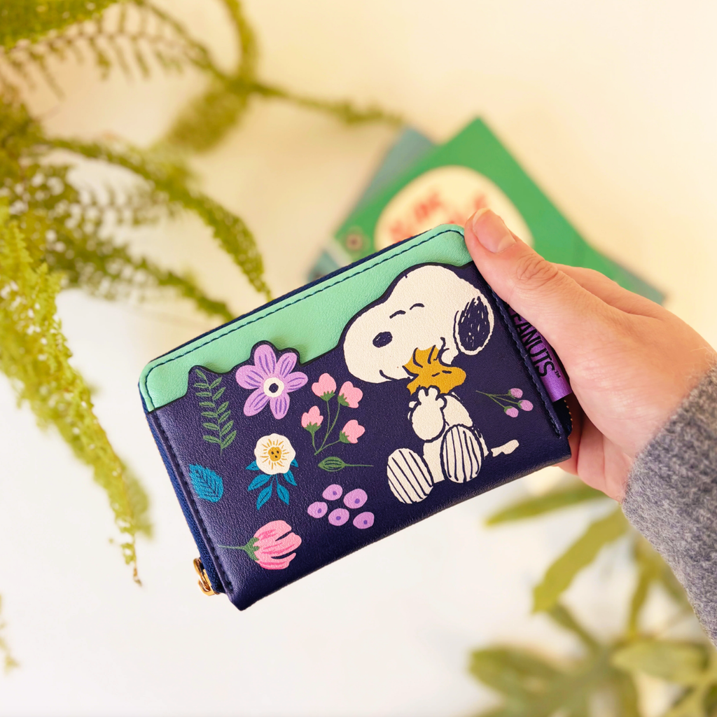 A hand holding a Peanuts-themed zip-around wallet featuring Snoopy and Woodstock on a dark navy background with colorful floral details, set against a softly blurred background with green foliage.