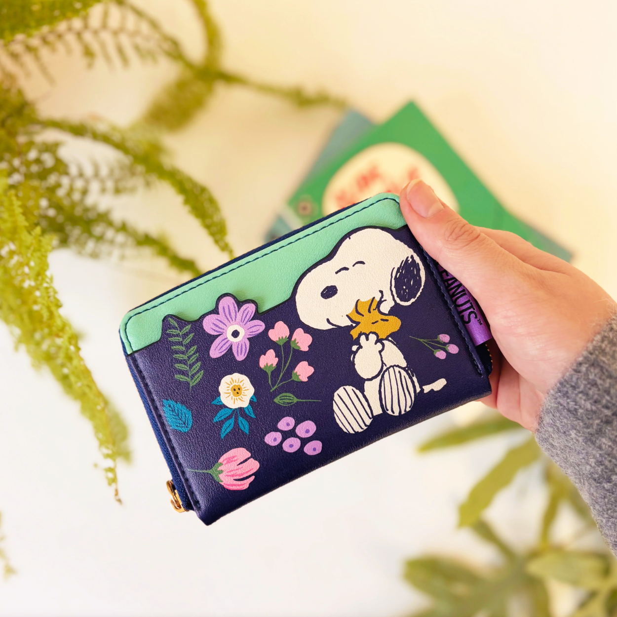 A hand holding a Peanuts-themed zip-around wallet featuring Snoopy and Woodstock on a dark navy background with colorful floral details, set against a softly blurred background with green foliage.