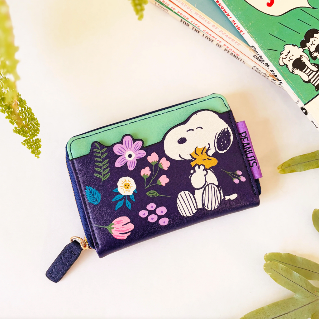 A Peanuts-themed zip-around wallet featuring Snoopy and Woodstock on a dark purple background with colorful floral illustrations, styled on a white surface with decorative leaves and a book.