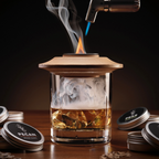 A clear glass tumbler with a light wood lid sits on a wooden table, filled with amber liquid and ice, with smoke rising from the top. A torch lighter hovers above the rim, and several round metal tins labeled PECAM are placed around it against a dark background.