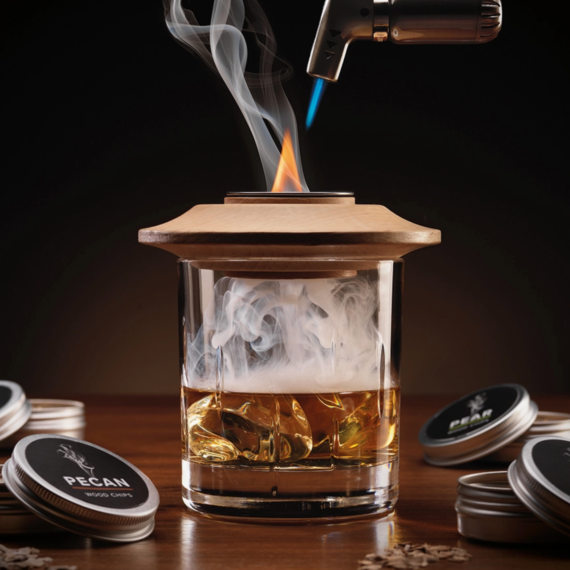 A clear glass tumbler with a light wood lid sits on a wooden table, filled with amber liquid and ice, with smoke rising from the top. A torch lighter hovers above the rim, and several round metal tins labeled PECAM are placed around it against a dark background.