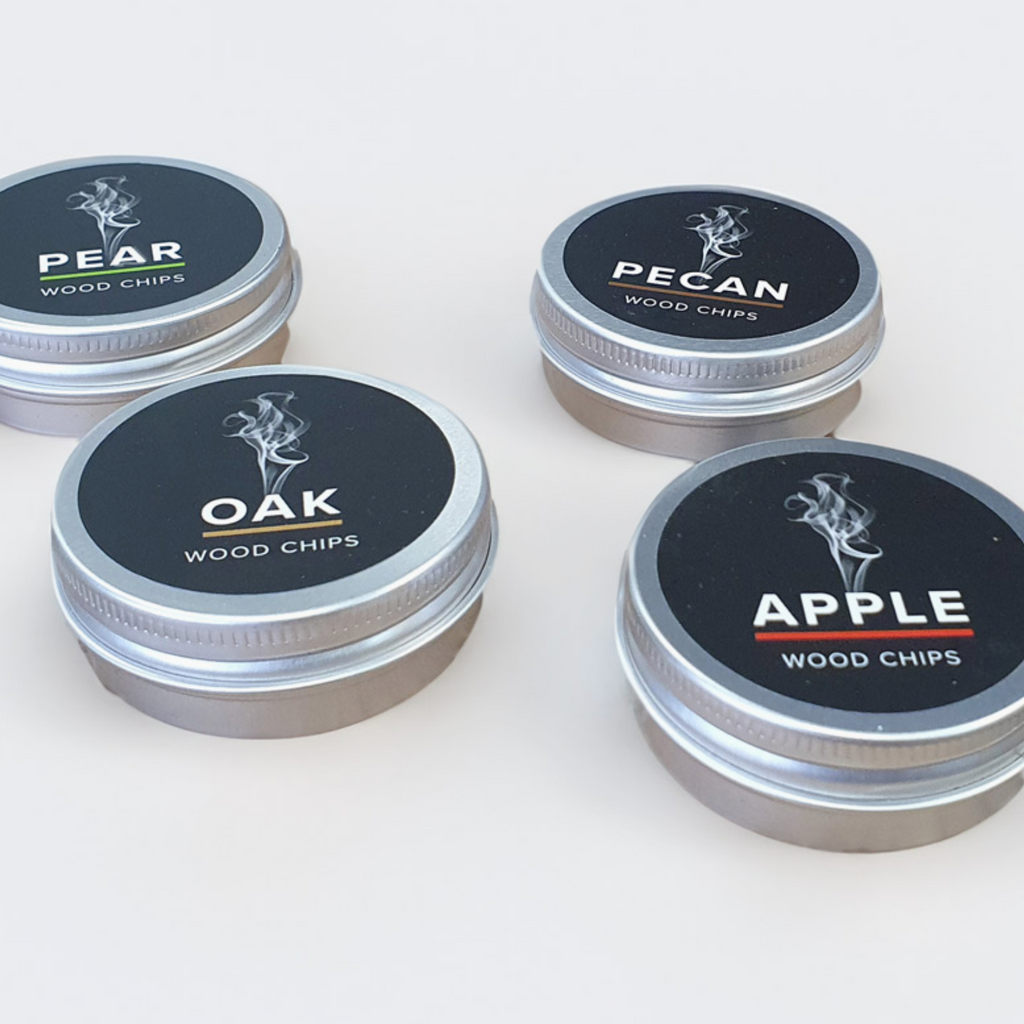 Four small round metal tins of wood chips arranged on a light neutral background. The lids are black with white text reading PEAR, PECAN, OAK, and APPLE, plus small flavor-specific accent lines.