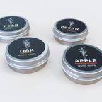 Four small round metal tins of wood chips arranged on a light neutral background. The lids are black with white text reading PEAR, PECAN, OAK, and APPLE, plus small flavor-specific accent lines.