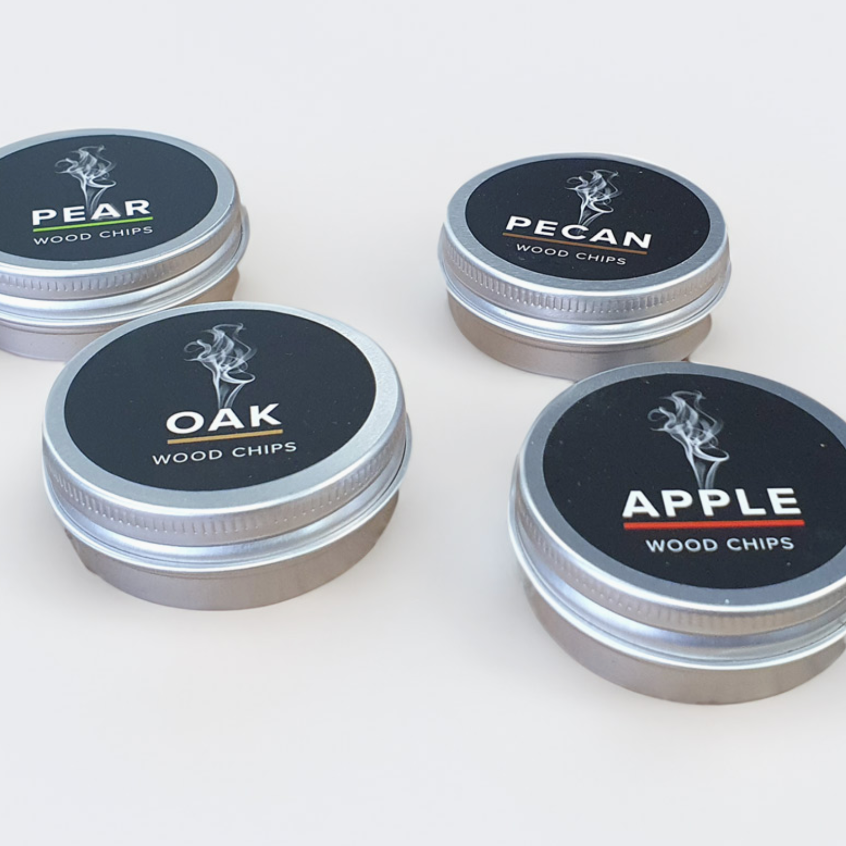 Four small round metal tins of wood chips arranged on a light neutral background. The lids are black with white text reading PEAR, PECAN, OAK, and APPLE, plus small flavor-specific accent lines.