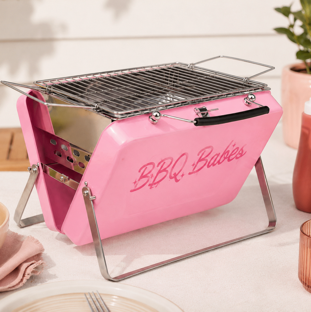 A pink portable mini grill with silver metal legs and a black handle, featuring the words 'BBQ Babies' in pink script on the front. The grill is styled on a table with condiments, a bowl of salad, and a plant in the background.