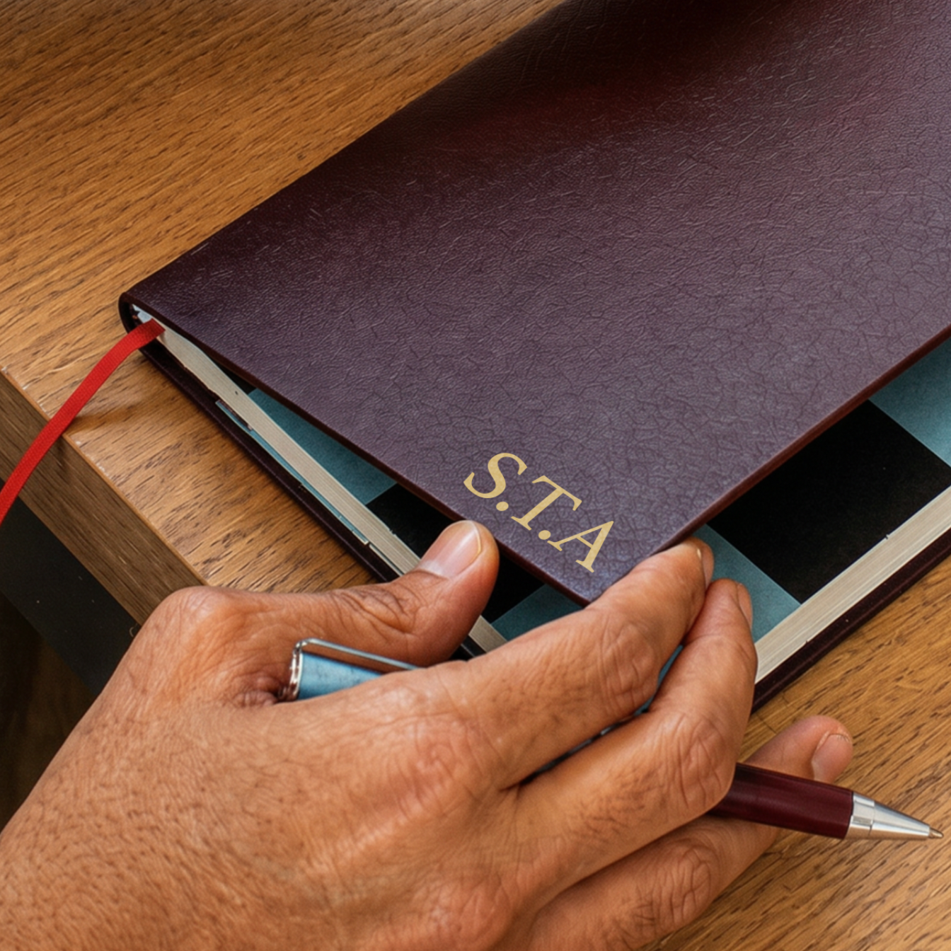 A close-up of a hand holding a pen over a dark brown leather-bound notebook with gold 'S.T.