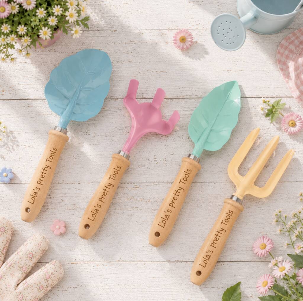 Four personalized gardening tools with wooden handles and colorful plastic heads are arranged on a white wooden surface with small flowers, gloves, a watering can, and daisies. The tools include a blue leaf-shaped trowel, a pink three-prong cultivator, a mint green leaf-shaped trowel, and a pale yellow three-prong cultivator.
