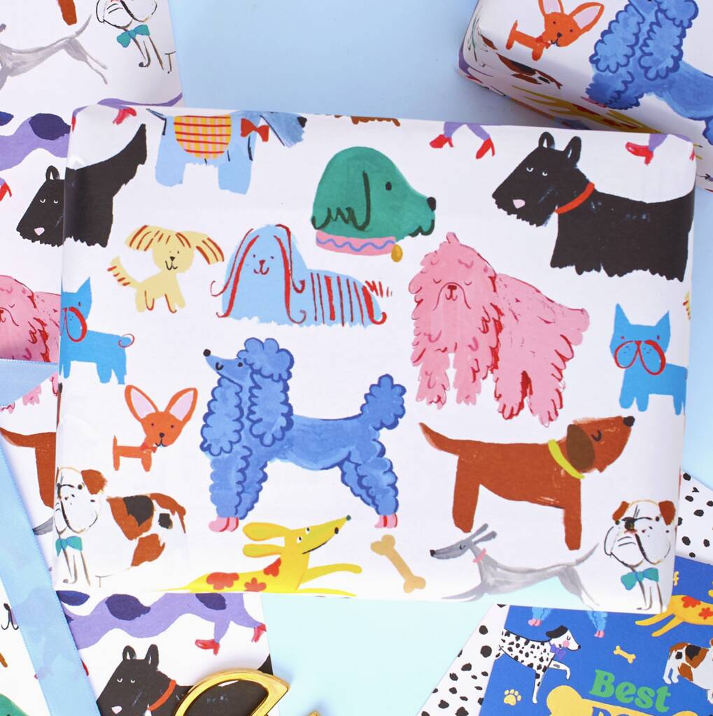 A close-up of a white gift wrap or fabric featuring a playful, colorful dog pattern with various dog breeds in blue, pink, black, brown, yellow, and green, styled on a light blue background.