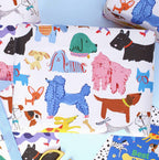 A close-up of a white gift wrap or fabric featuring a playful, colorful dog pattern with various dog breeds in blue, pink, black, brown, yellow, and green, styled on a light blue background.