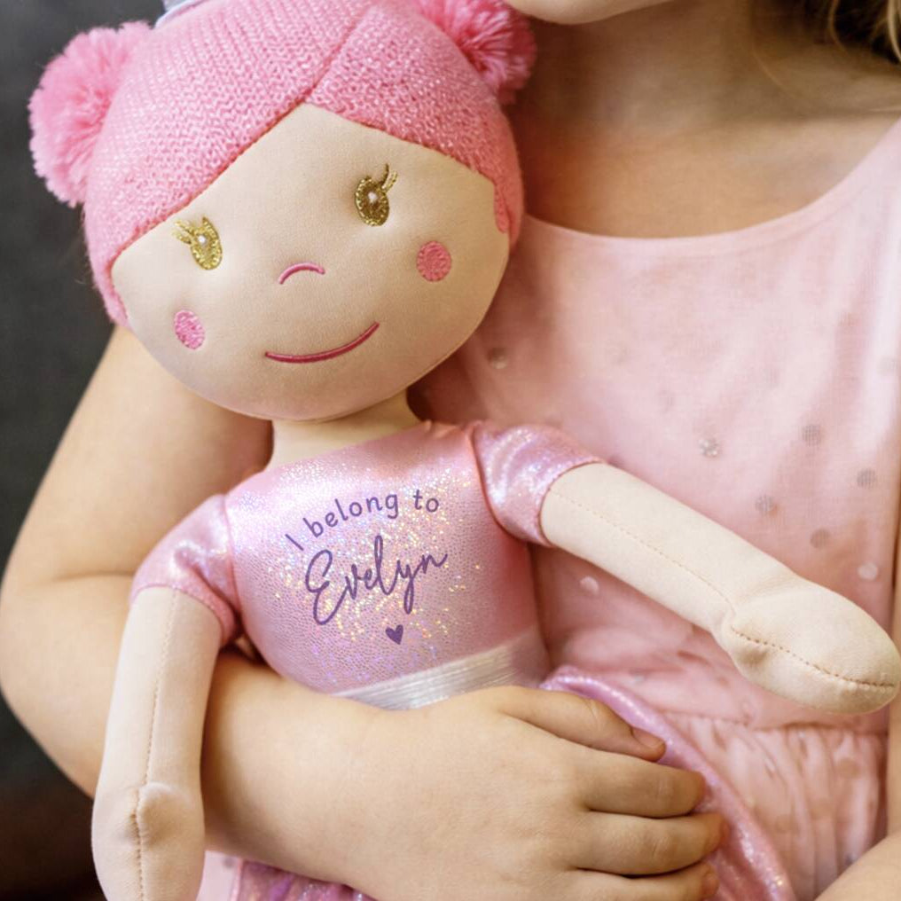 A close-up lifestyle shot of a child in a pink dress holding a custom embroidered plush doll. The doll has a pink knit hat with pom-poms, a beige face, and a glittery pink shirt that reads 'I belong to Evelyn'.