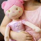 A close-up lifestyle shot of a child in a pink dress holding a custom embroidered plush doll. The doll has a pink knit hat with pom-poms, a beige face, and a glittery pink shirt that reads 'I belong to Evelyn'.
