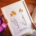 A framed custom flower print held by a child, featuring two gold flower petals with black line art and the text 'Daddy's Flowers' on a white background.