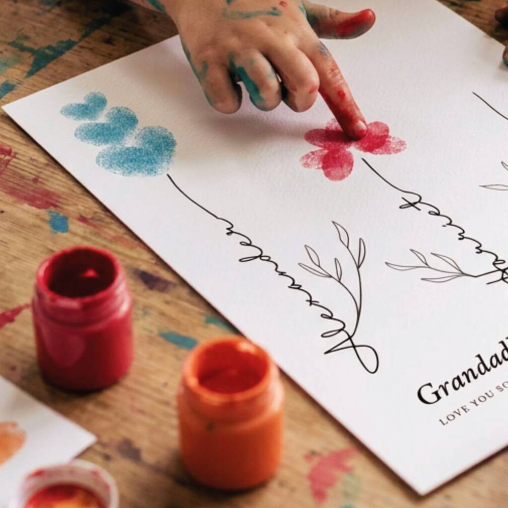 A close-up of a child's hand with colorful paint stains pressing a red paint dot onto a white art print, with two open paint pots on a wooden table.